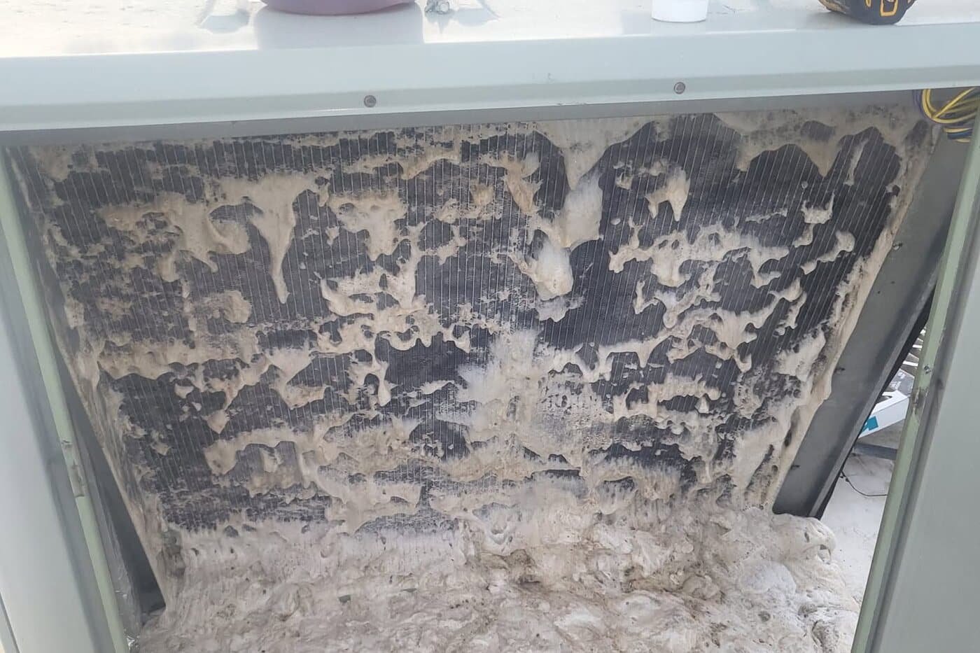 An open equipment cabinet with the coil face packed solid behind thick matted grey debris.
