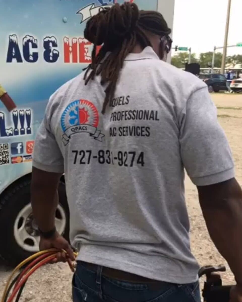 A Quel's Professional AC technician at the back of the service truck in a company shirt, carrying refrigerant hoses.