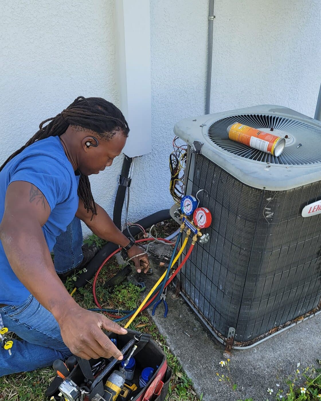 A technician crouched at an outdoor condenser with a refrigerant manifold and gauges connected.